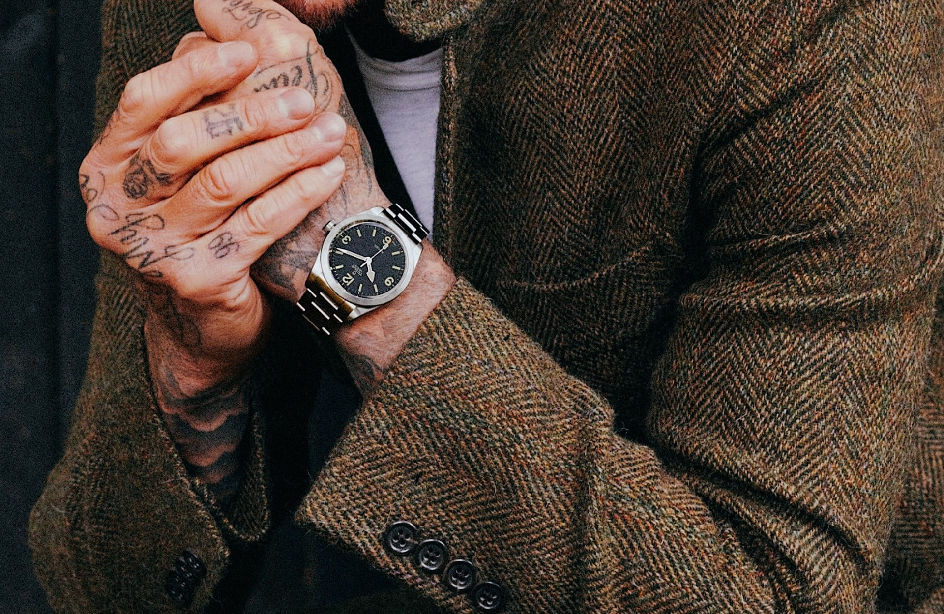 Steel field watch on a bracelet, worn under a brown herringbone tweed jacket